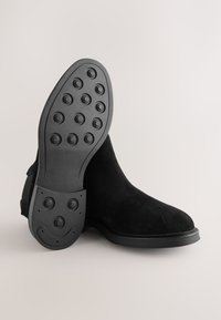 Melnas zamšādas potītes zābaki ar gludu apdari, ovālu gumijas zolīti ar aprindu vilkšanas rakstiem un elegantu siluetu.