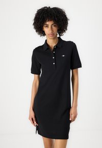 Rochie polo neagră din bumbac, cu guler, mâneci scurte și un accent subtil cu logo. Fantele laterale îmbunătățesc mobilitatea.