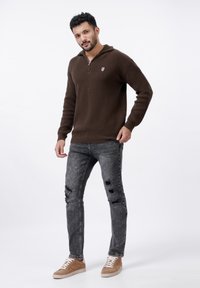 Brun stickad tröja med dragkedja i kragen och emblem, kombinerad med slitna gråjeans och bruna sneakers med vita snören.