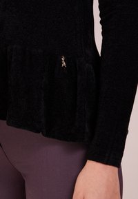 Zwarte, pluche top met lange mouwen en een gerimpelde zoom, met een gouden hanger detail bij de taille. De textuur lijkt zacht en warm, geschikt voor casual wear.