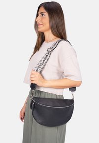 Schwarze Leder-Crossbody-Tasche mit einer strukturierten Oberfläche, ausgestattet mit einem silbernen Reißverschluss und einem gemusterten, verstellbaren Tragegurt in Schwarz und Weiß.