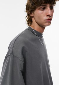 Sweatshirt gris oversize avec un col rond, des épaules tombantes et des détails de couture visibles. Tissu doux et texturé avec une coupe décontractée.