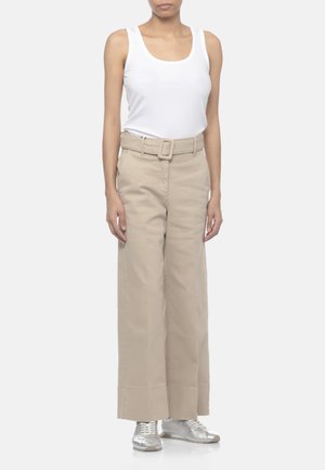 Donna con canottiera bianca, pantaloni beige a gamba larga con cintura e sneakers argento, in piedi davanti a uno sfondo semplice.
