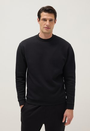 Hombre con cabello oscuro que lleva una sudadera negra lisa de manga larga y pantalones negros a juego, de pie contra un fondo claro.