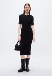 Μαύρο μίντι φόρεμα με ριμπ ύφανση, κοντά μανίκια και λεπτομέρειες με κουμπιά, συνδυασμένο με μικρή μαύρη τσάντα χειρός και μαύρες μπότες μέχρι το γόνατο.