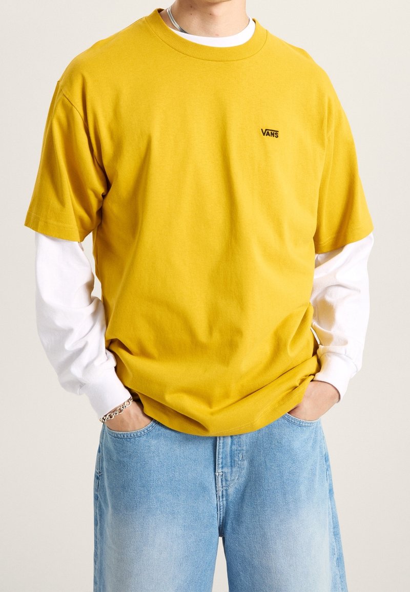 Personne portant un t-shirt Vans jaune par-dessus une chemise blanche à manches longues avec un jean bleu clair, les mains dans les poches, sur un fond uni.