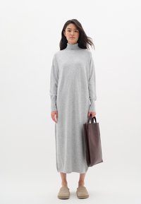 Grå, långärmad, polokragsklänning i ett mjukt tyg med en lös passform. En mörkbrun tote-väska hålls i ena handen och beige tofflor används.