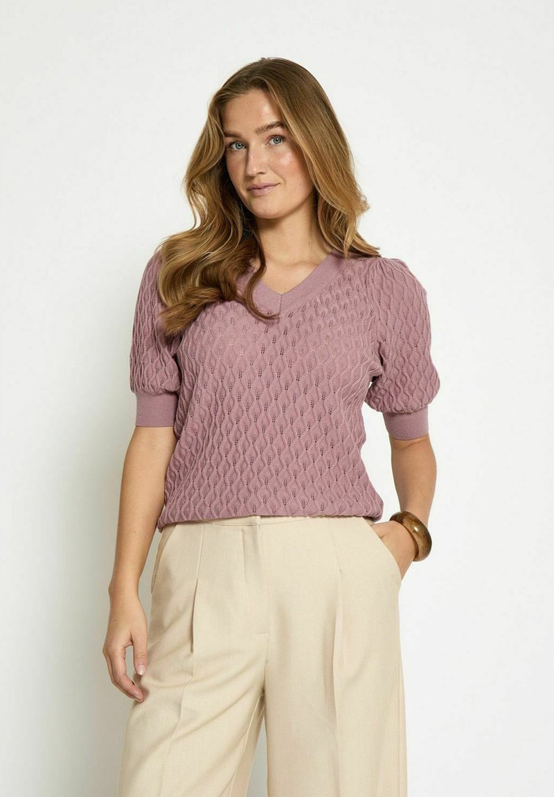 Knitwear lyhythihainen mauve-väri, jossa on teksturoitu timanttikuvio ja V-nuora, yhdistettynä vaaleanbeigeihin housuihin.