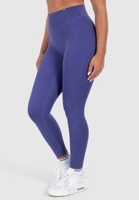 Leggings viola a vita alta realizzati in tessuto elastico, caratterizzati da una texture liscia e senza cuciture visibili, abbinati a scarpe da ginnastica bianche.