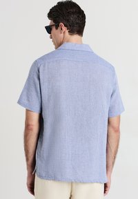 Uomo che indossa una camicia a maniche corte texturizzata di colore azzurro chiaro e pantaloni beige, rivolto di spalle, con occhiali da sole neri su uno sfondo chiaro e semplice.