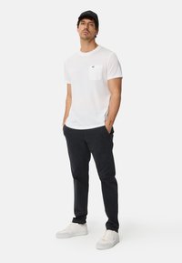 White cotton t-shirt with a chest pocket, paired with dark navy trousers and white sneakers. Model stands with hands in pockets, wearing a black cap.