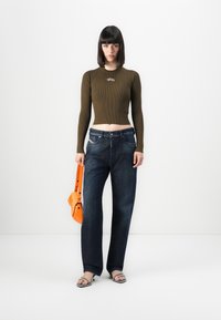 Ženska stoji pred belo ozadjem, oblečena v skrajšano olivno pulover, ohlapne temno jeans hlače, sandale, v eni roki drži oranžno torbico.