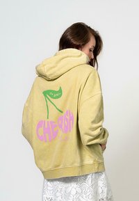Žltá bavlnená mikina s oversized strihom, na prednej strane s grafickým znázornením čerešní a slovom "CHERISH" v ružovej farbe, a na zadnej strane so zeleným bannerom s nápisom "OH APRIL".