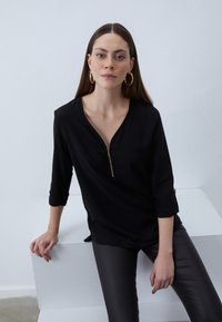 Black top with a gold zipper at the neckline, three-quarter sleeves, and a relaxed fit. Model wears shiny black leggings.