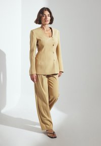 Costume beige sur mesure comprenant un blazer ajusté avec un décolleté carré, trois boutons et un pantalon large ; tissu doux et coupe décontractée.