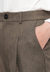 Hand in de zak van bruin gestructureerde broek gedragen met een ingestopt wit overhemd, met zwarte knoop en riemlusdetails.