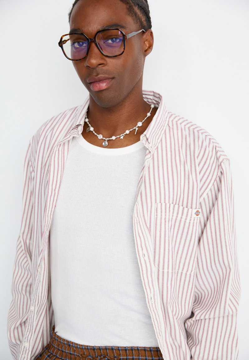 Gestreiftes Hemd in Pastellrosa und Weiß über einem weißen ribbed Tanktop. Runde Halskette mit silbernen Charms und blaugemäßen Brille.
