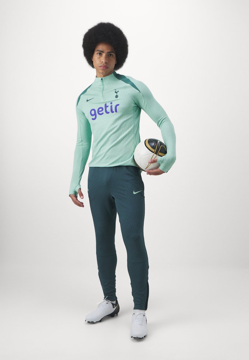 Haut de sport vert menthe à manches longues et col zippé, pantalon vert foncé et chaussures de football blanches. Tient un ballon de football à motifs.
