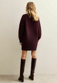 Abito maglione oversize borgogna con maniche lunghe, tessuto testurizzato e orlo che cade sopra il ginocchio, abbinato a stivali lucidi marroni alti fino al ginocchio.
