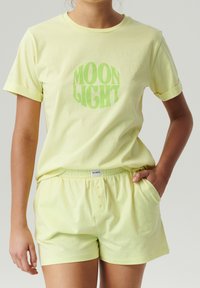 Jasnożółta bawełniana koszulka z grafiką "MOON LIGHT" w zielonym kolorze. Pasujące szorty z elastycznym wstążką w talii i dwiema przednimi kieszeniami, również jasnożółte.