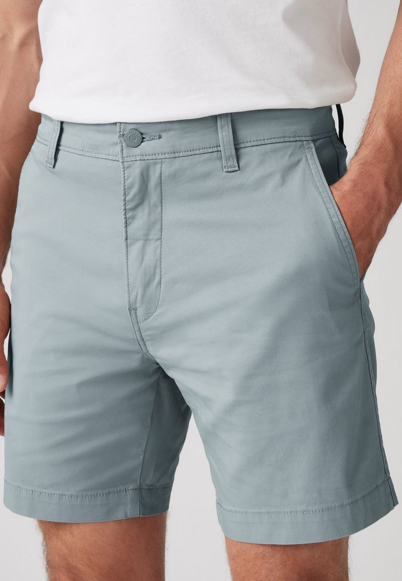 Shorts en coton gris clair avec un devant plat, des poches latérales et une fermeture à bouton. Texture lisse et détails de couture soignés le long de l'ourlet.