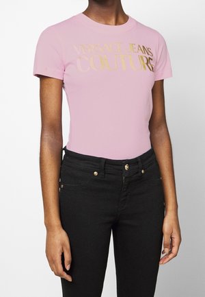 Persoon draagt een roze Versace Jeans Couture t-shirt met gouden letters en een zwarte hoge taille jeans met gouden knopen.