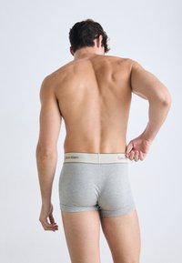 Een shirtloze man die de tailleband van zijn lichtgrijze Calvin Klein boxershort aanpast, staand tegen een effen witte achtergrond.