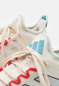 Jasnobeżowe buty sportowe z czerwonymi i niebieskimi akcentami. Siatkowa cholewka, teksturowany materiał i warstwowy design z trzema niebieskimi paskami na pięcie.