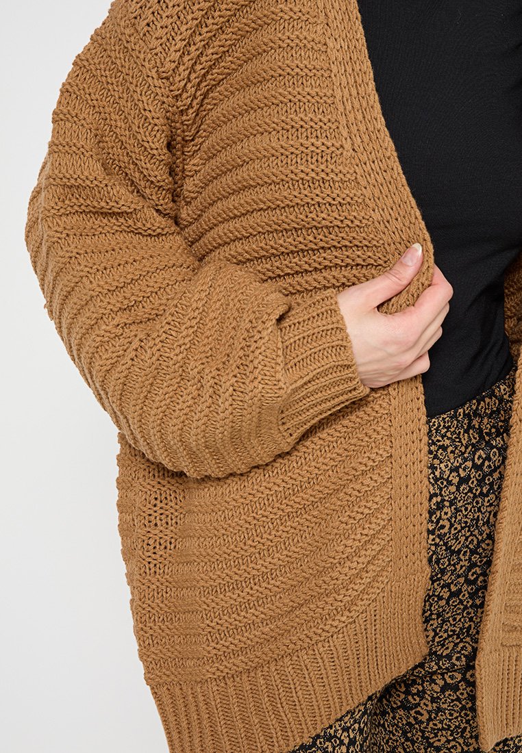 Sweter z dzianiny w ciepłym brązowym kolorze, z fakturowanym, żebrowanym wzorem, szerokimi rękawami i luźnym krojem, pasującym do wzorzystych spodni.