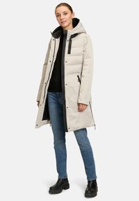 Parka beige clair matelassée avec capuche amovible, fermeture éclair avant et accents noirs. Portée sur un haut noir et un jean bleu avec des bottes chelsea.