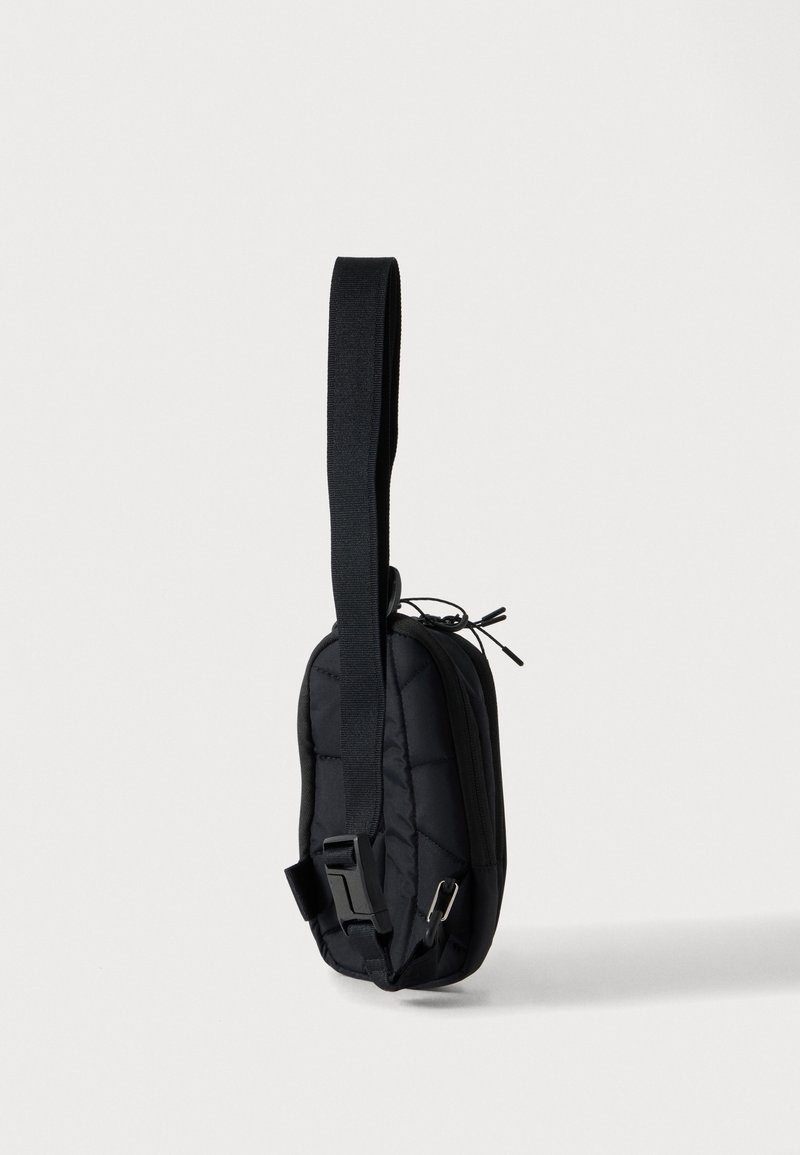 Lille sort crossbody- eller bæltetaske med justerbar rem og lynlåslukning, vist hængende lodret mod en ensfarvet hvid baggrund.