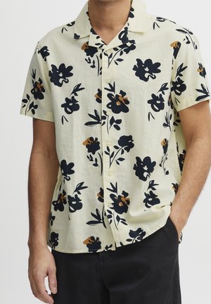 Mann trägt ein beigefarbenes Kurzarm-Hemd mit schwarzen und braunen Blumenmustern, kombiniert mit schwarzer Hose und einer Hand in der Tasche.