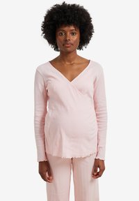 Woman with natural curly hair wearing a light pink ribbed long sleeve top and matching pants, standing against a white background.