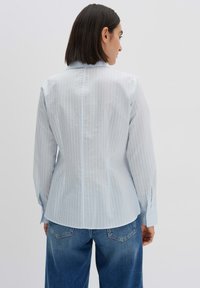Camicia a righe blu chiaro con colletto, maniche lunghe e orlo arrotondato. Il tessuto appare liscio con una leggera lucentezza. Indossata con jeans di denim.