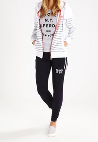 Haut zippé blanc avec des accents orange, t-shirt gris à rayures, et joggers bleu marine avec un détail de logo blanc et des rayures sur les côtés. Look décontracté de vêtements de sport.