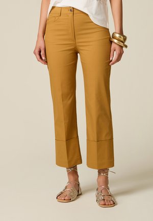 Donna con pantaloni larghi color senape, risvoltati alla caviglia, abbinati a sandali bassi con cinturini e braccialetti dorati a un polso.