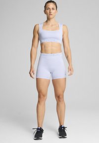 Sujetador deportivo de color púrpura claro y pantalones cortos a juego, hechos de material elástico, con un logo de PUMA en la cintura. Unos zapatos deportivos negros completan el look.