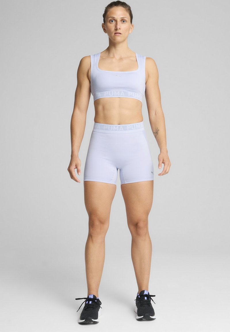 Sujetador deportivo de color púrpura claro y pantalones cortos a juego, hechos de material elástico, con un logo de PUMA en la cintura. Unos zapatos deportivos negros completan el look.