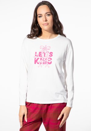 Frau mit langen dunklen Haaren trägt ein weißes Langarmshirt mit pinkem "LET'S KISS" Text und floralem Design, kombiniert mit roten karierten Hosen.