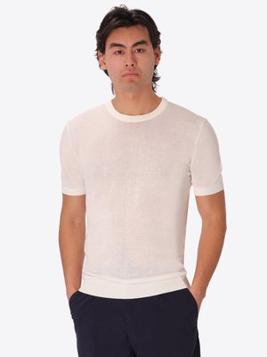 Uomo con capelli neri di media lunghezza che indossa una maglia bianca a maniche corte e pantaloni scuri con le mani in tasca.