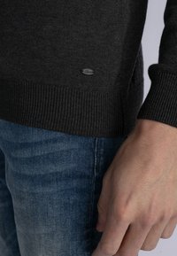 Dark gray knit sweater with ribbed cuffs, featuring a small branded tag on the lower side. Paired with blue denim jeans.