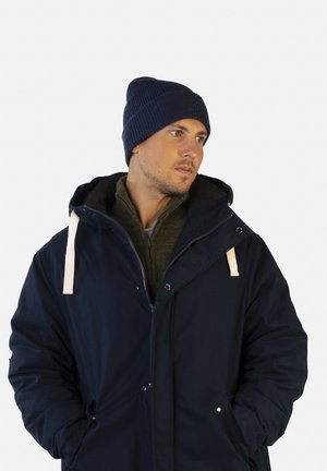 Parka bleu marine avec cordons blancs, dotée d'une fermeture éclair et de poches latérales. Portée par-dessus un pull en tricot vert foncé et un bonnet bleu marine assorti.