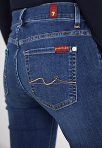 Dark blue denim jeans with a fitted design, featuring a red tag labeled '7' and a decorative golden stitching pattern on the back pocket.