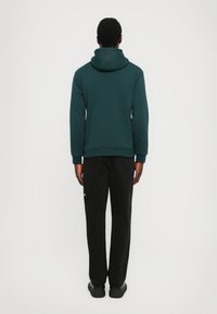 Felpa con cappuccio di un verde scuro, dalla vestibilità comoda e con polsini a costine, abbinata a pantaloni neri e scarpe nere. Design semplice, dettagli minimi.