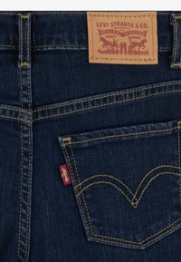 Vaqueros de mezclilla azul oscuro con una etiqueta de cuero que dice "Levi Strauss & Co." y una etiqueta roja cerca del bolsillo trasero, acentuados con costuras amarillas.