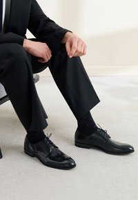 Chaussures habillées en cuir noir avec une finition brillante, un design à lacets et un bout arrondi. Associées à des chaussettes sombres avec des motifs subtils.