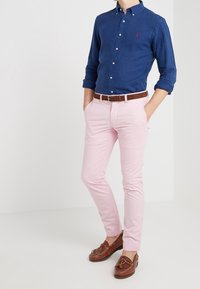 Blå skjorta med knappar, rosa byxor, brunt läderbälte och bruna loafers. Skjortan har en liten logotyp; byxorna är slim-fit.