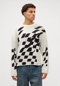 Strikkegenser i kremfarge med et dristig svart geometrisk mønster. Har rund hals, lowered skuldre og en avslappet passform.