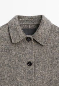 Cappotto grigio in misto lana con colletto, dotato di chiusura a bottone nera e una texture sottile. L'etichetta è visibile all'interno del colletto.