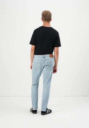 Homme debout de dos portant un t-shirt noir, un jean bleu clair et des baskets noires avec des bandes blanches sur un fond blanc.